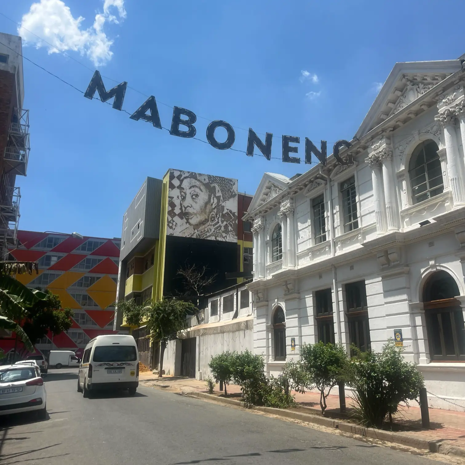 Maboneng Precinct street in Johannesburg with overhead MABONENG sign and mural on a building under blue sky