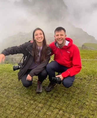 Ryan and Sara of Across Every Border crouching on a grassy hill with cameras against misty Machu Picchu ruins