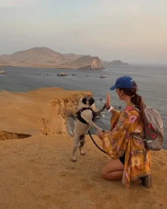 Travel creator Lora giving a high-five to her dog Puglo on a coastal cliff overlooking rocky islands and the ocean