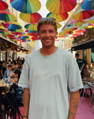 Content creator Marko smiling on a colorful street lined with hanging umbrellas at an outdoor café