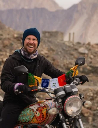 Travel blogger Will Hatton laughing on a decorated motorcycle in a rugged mountain landscape