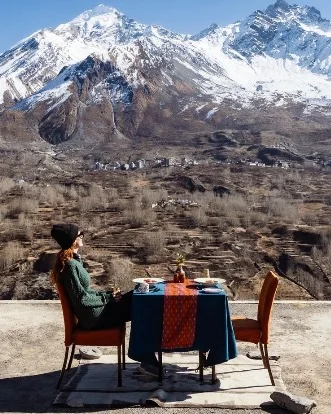 Travel writer Yvette Bax dining outdoors with snow-capped mountains and a valley village in the background