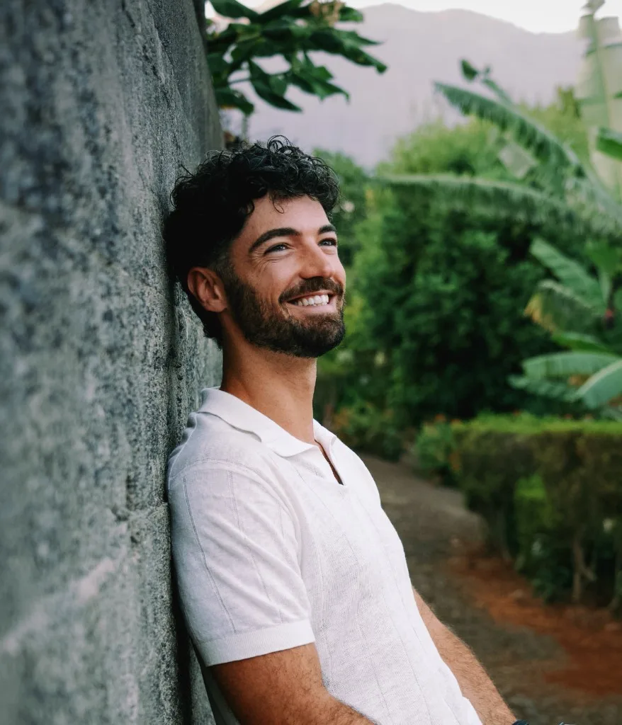 Henry Snow smiling outdoors beside a stone wall in a tropical garden