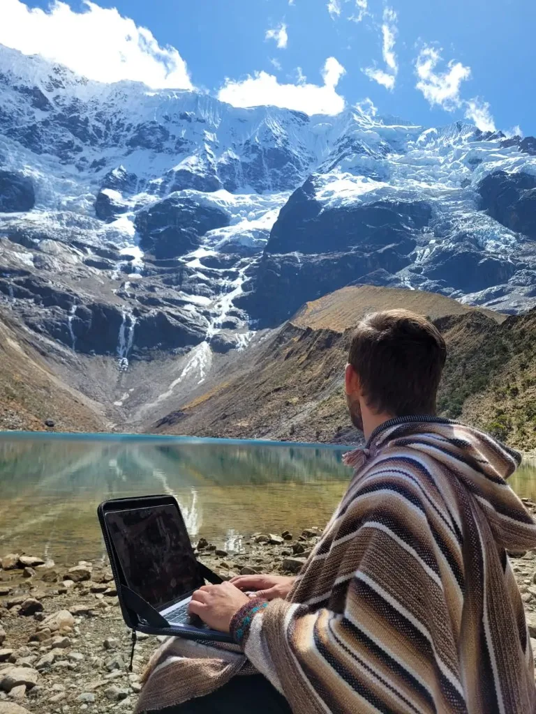 Igor Kondrat working on a laptop on a rocky shore beside a turquoise glacial lake and snow-capped mountains