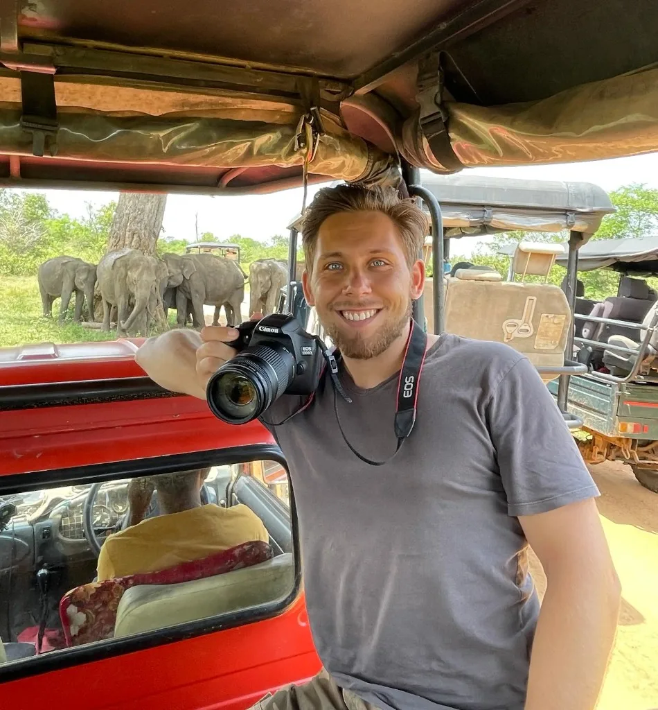 Travel journalist Igor Kondrat holding a Canon camera beside a safari jeep with elephants in the background
