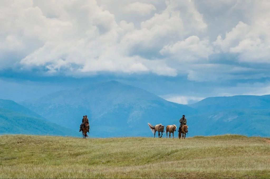 Horse riders on the Mongolian steppe with pack horses, rolling grassland, and distant blue mountains under a wide sky